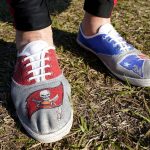 
              FILE - A fan arrives wearing shoes from both Tampa Bay Buccaneers quarterback Tom Brady's teams before the NFL Super Bowl 55 football game between the Kansas City Chiefs and Tampa Bay Buccaneers in Tampa, Fla., in this Sunday, Feb. 7, 2021, file photo. Brady hasn't played an NFL football game in New England since Jan. 4, 2020. But remnants from his 20-year run with the Patriots remain everywhere in Foxborough. With Brady's new Tampa home just as rabid about the player who brought it a championship in Year 1, it's created a tug-of-war between the fan bases as he gets set to return to the place where his career began. (AP Photo/Mark Humphrey, File)
            