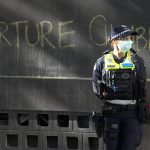 
              A police officer stands by a slogan on the wall outside the Park Hotel, used as an immigration detention hotel where tennis player Novak Djokovic is confined in Melbourne, Australia, Sunday, Jan. 9, 2022. After five nights in hotel detention Djokovic will get his day in court on Monday, Jan. 10, in a controversial immigration case that has polarized opinions in the tennis world and elicited heartfelt support for the star back home in his native Serbia. (AP Photo/Mark Baker)
            