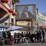 
              FILE - Residents line up at a security checkpoint into the Hotan Bazaar where a screen shows Chinese President Xi Jinping in Hotan in western China's Xinjiang region on Nov. 3, 2017. The Chinese president, hosting a Winter Olympics beleaguered by complaints about human rights abuses, has upended tradition to restore strongman rule in China and tighten Communist Party control over the economy and society. (AP Photo/Ng Han Guan, File)
            