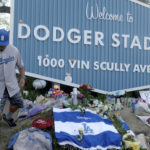 
              Los Angeles Dodger fans, including Rudy Escobar with his son Rodolofo, 7, visit a memorial for Los Angeles Dodgers broadcaster Vin Scully at the entrance to Dodger Stadium on Wednesday, Aug. 3, 2022, in Los Angeles. The Hall of Fame broadcaster, whose dulcet tones provided the soundtrack of summer while entertaining and informing Dodgers fans in Brooklyn and Los Angeles for 67 years, died Tuesday night. He was 94. (Dean Musgrove/The Orange County Register via AP)
            