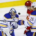 
              Buffalo Sabres goalie Eric Comrie, left, deflects the puck as Calgary Flames forward Mikael Backlund, center, closes in during the third period of an NHL hockey game Thursday, Oct. 20, 2022, in Calgary, Alberta. (Jeff McIntosh/The Canadian Press via AP)
            